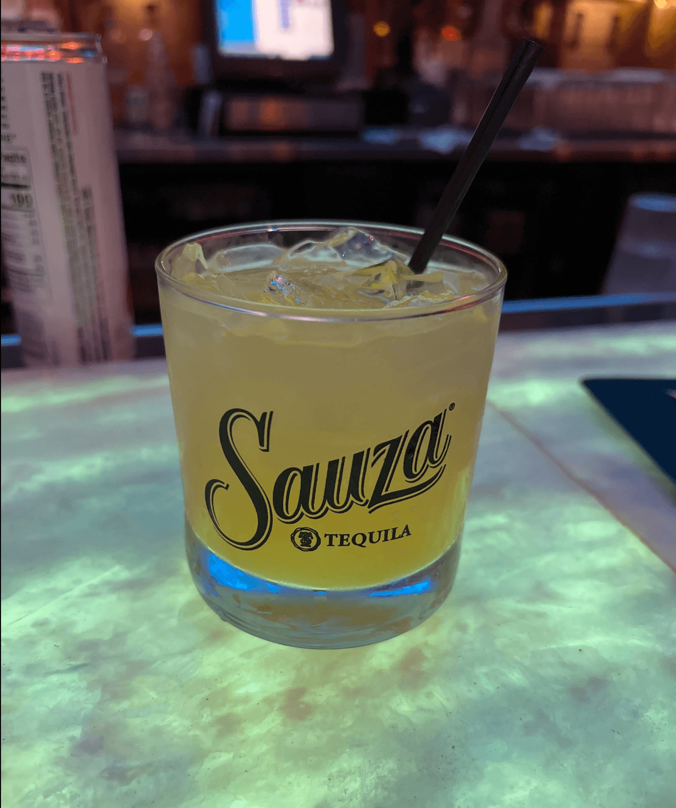Yellow cocktail with ice and straw in a Sauza Tequila glass on a glowing bar.