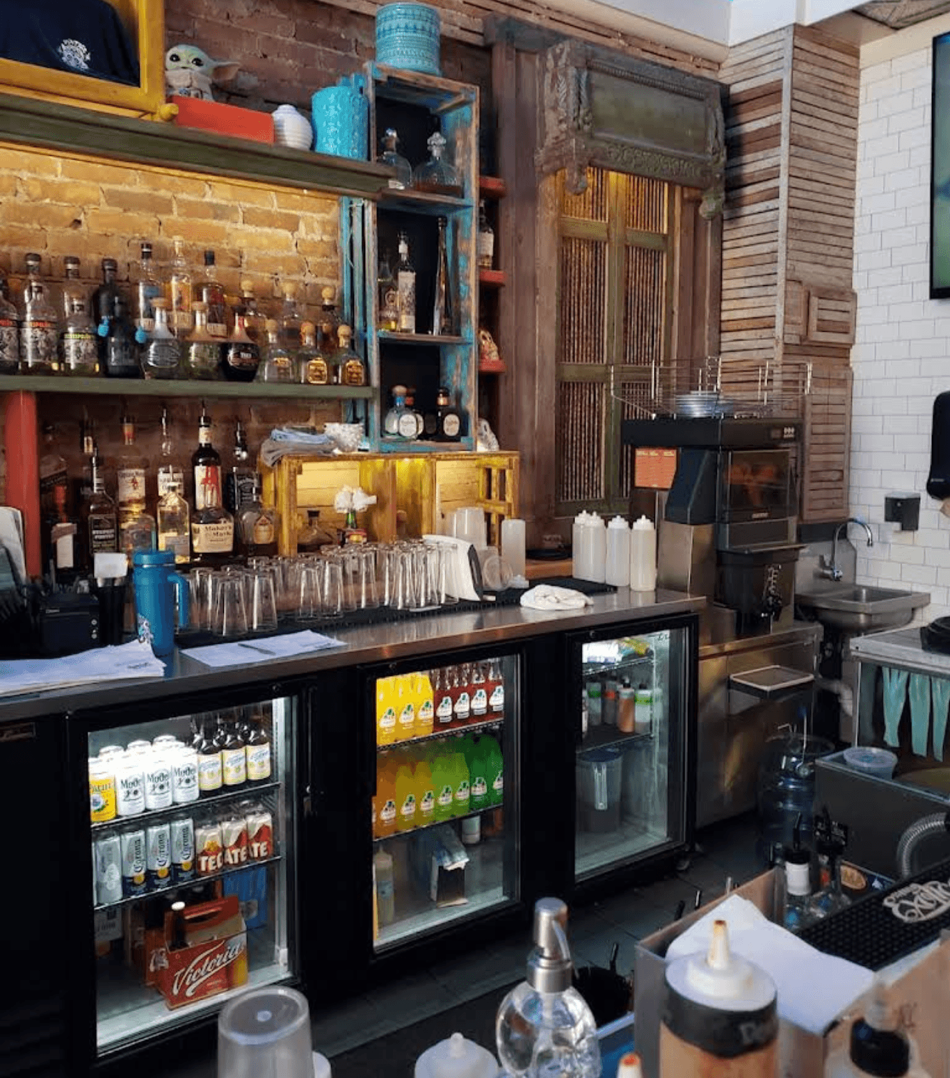 Bar counter with liquor bottles on brick shelves above refrigerators filled with assorted drinks.